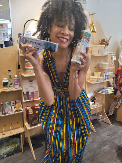 Lecretia Akines holding a bottle of Black Seed Herbal Toothpaste and a bottle of mouthwash, smiling in a retail environment.
